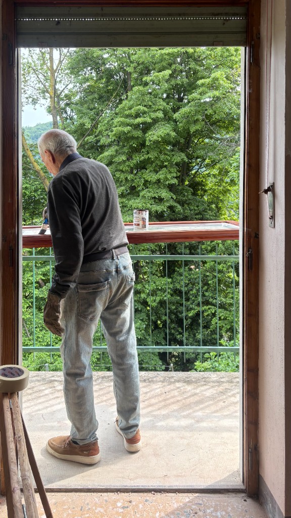 Uomo anziano che vernicia una cornice di legno posata sulla ringhiera del balcone, bosco sullo sfondo